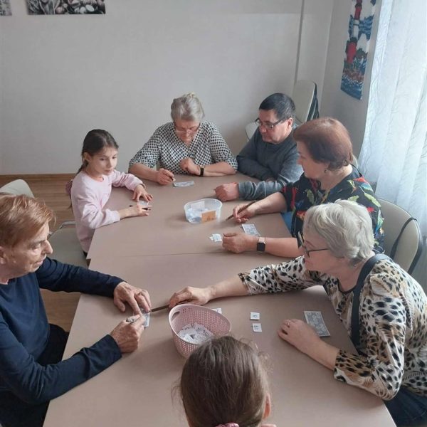 Przy dużym stole siedzą osoby dorosłe i dzieci, grają wspólnie w gry..