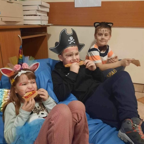 Troje dzieci siedzi na niebieskiej puffie. dzieci jedzą pizzę. Dziewczynka jest w stroju jednorożca, chłopcy w stroju pirata i kota.