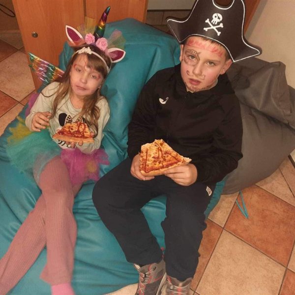 Dziewczynka i chłopiec siedzą na zielonej puffie. i jedzą pizzę. Chłopiec w stroju pirata, dziewczynka w stroju jednorożca.