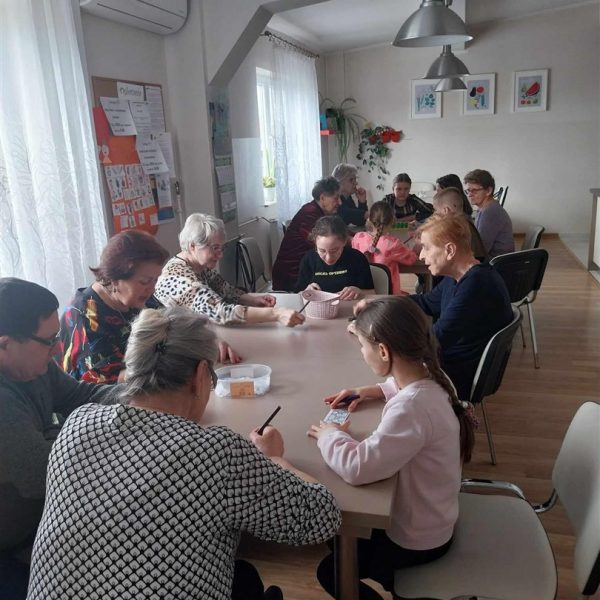 Kilkanaście osób siedzi przy dwóch stołach, są to osoby starsze i dzieci, grają w gry.